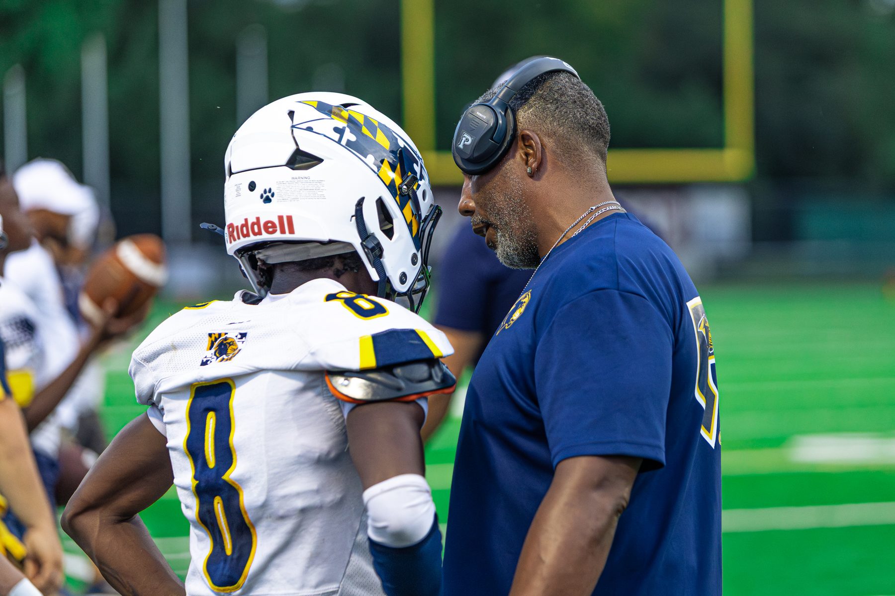 Football coach in a headset leaning in to talk with player number 8 on the sideline during a day game
