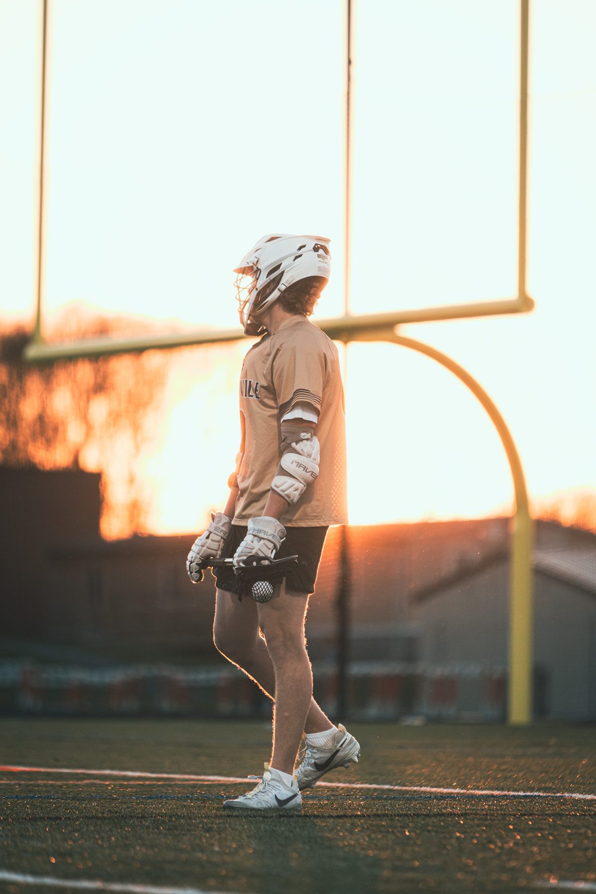 Lacrosse player walking across the field silhouetted by a low orange sun behind the goalpost