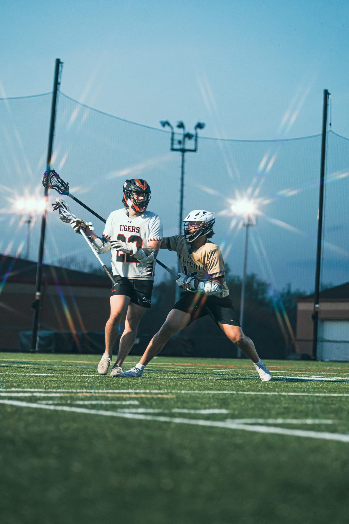Lacrosse defender pressuring a ball carrier at dusk beneath bright stadium lights