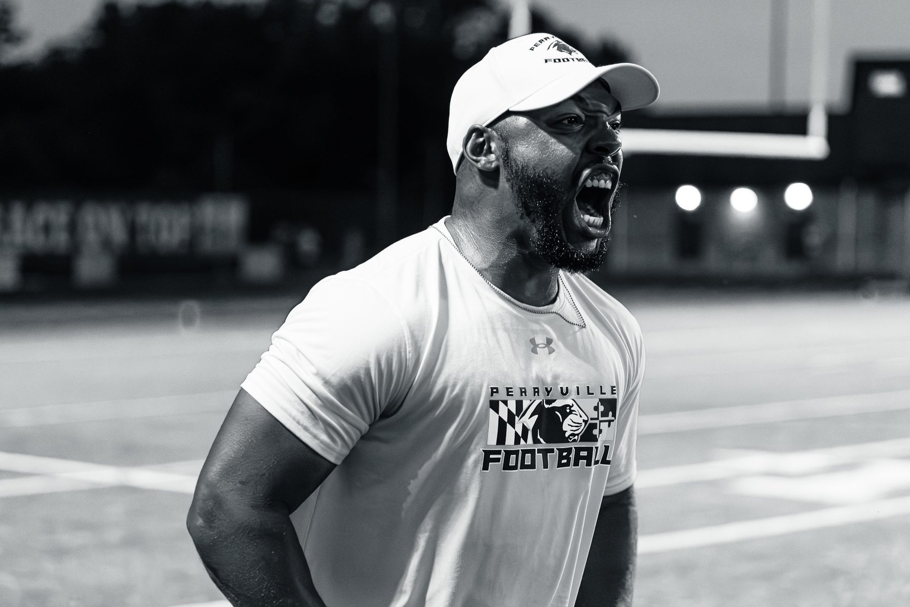 Black and white photo of a football coach shouting on the field at night with stadium lights blurred behind him